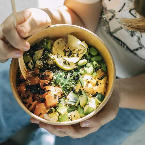 Person holding a poke bowl with salmon, avocado, edamame, and seaweed.