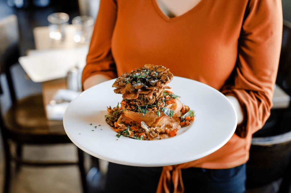 Person holding a white plate with a fancy, stacked gourmet dish.