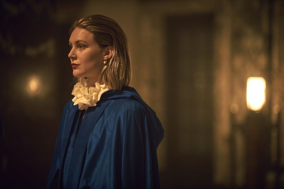 Person in a blue cloak with white ruffled collar, in a dimly lit, elegant room.