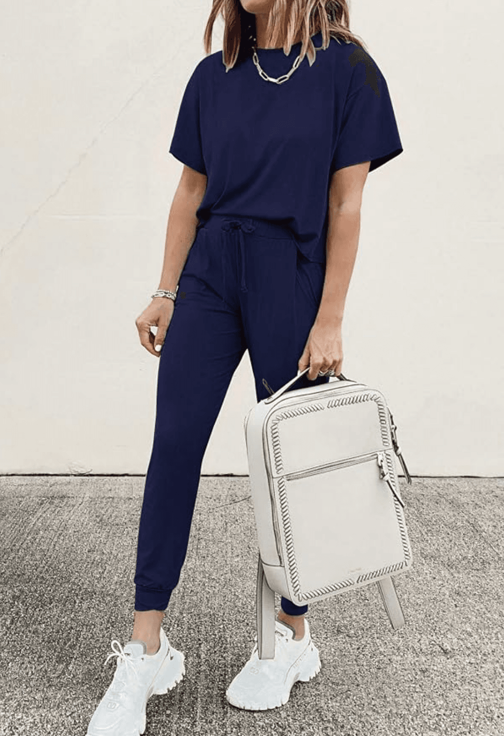 Person in a blue outfit holding a white backpack, wearing white sneakers.