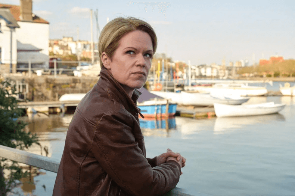 Person in a brown jacket gazing over a calm harbor with moored boats.