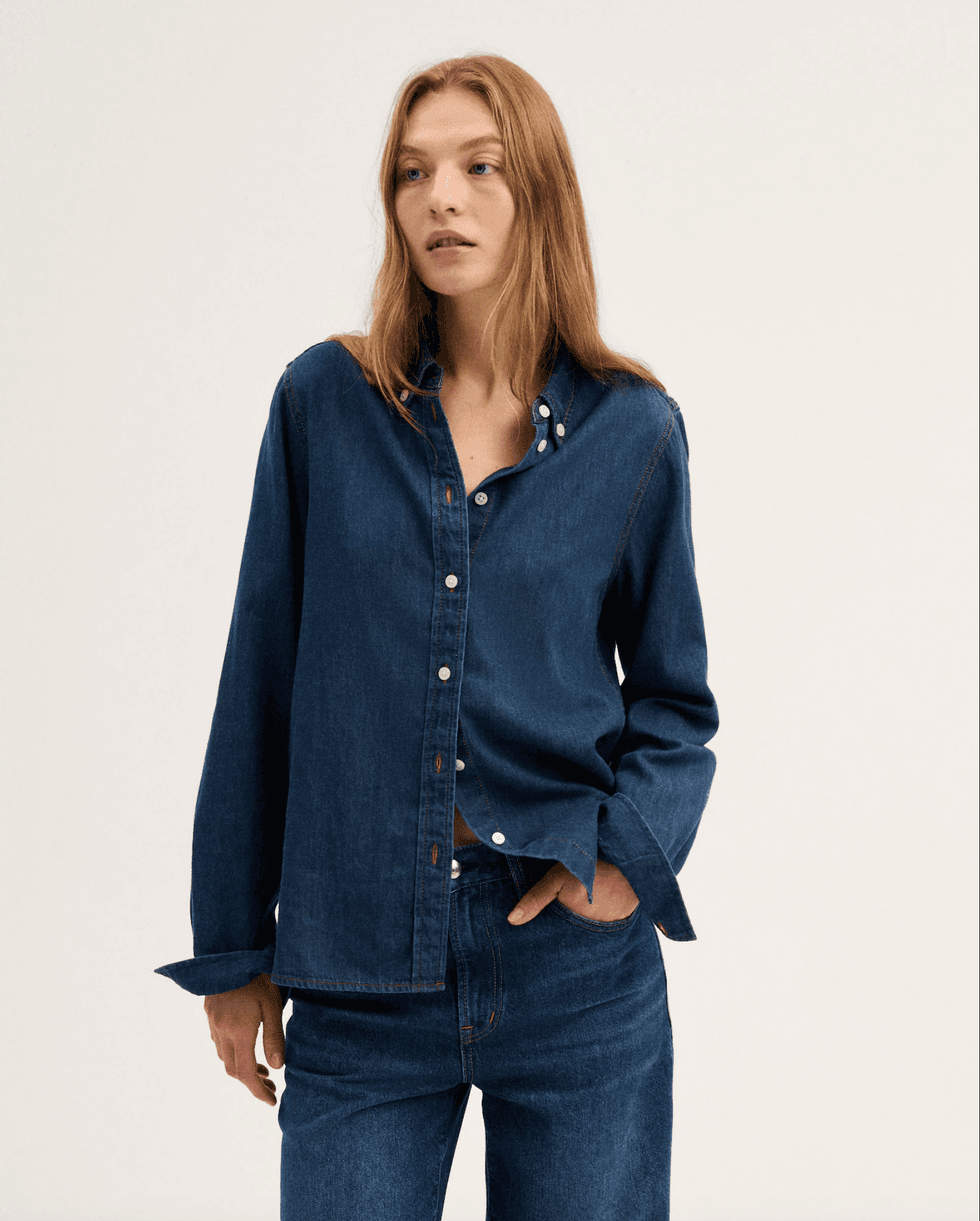 Person in a denim shirt and jeans against a plain background.