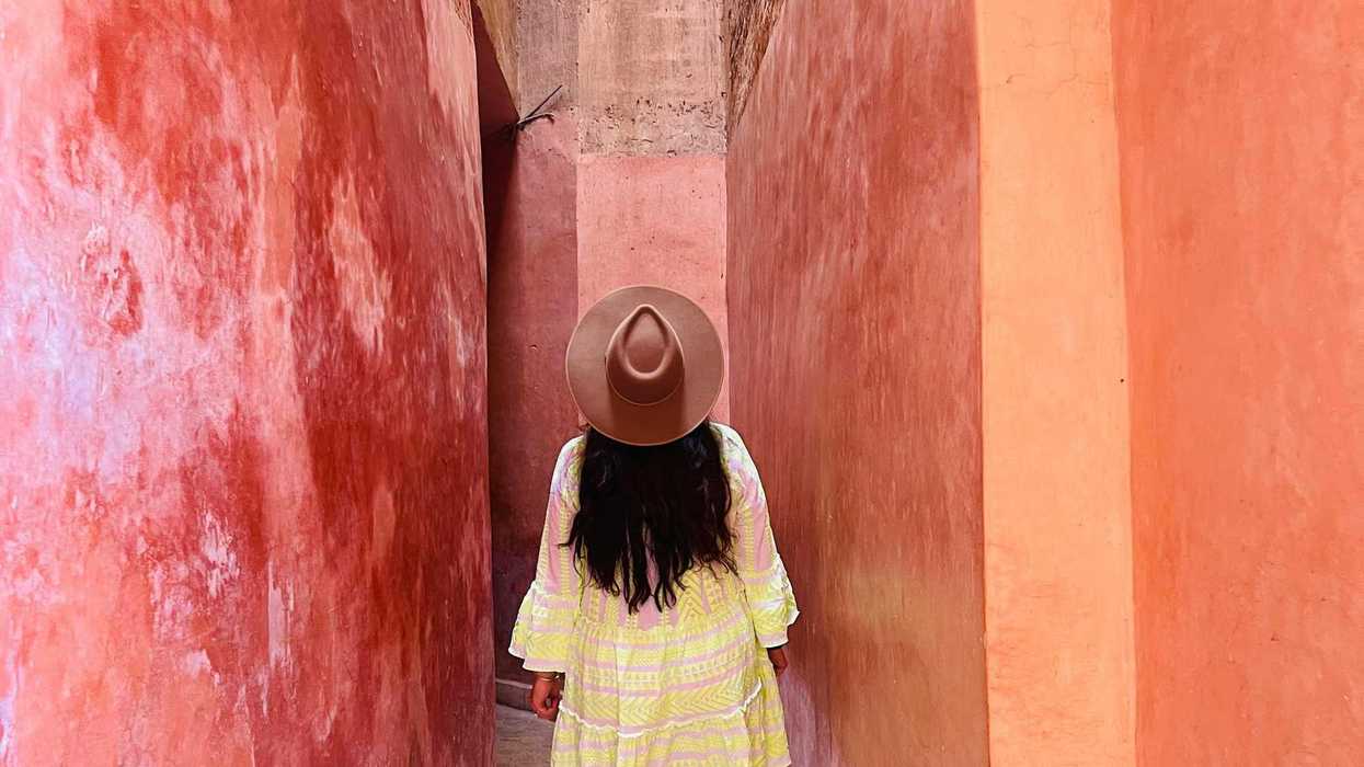 Person in a dress and hat walking down a narrow, sunlit, pink alley.