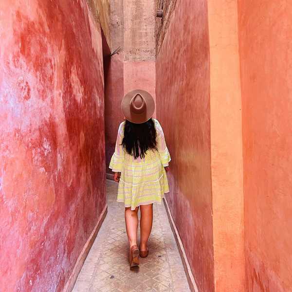 Person in a dress and hat walking down a narrow, sunlit, pink alley.