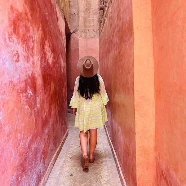 Person in a dress and hat walking down a narrow, sunlit, pink alley.