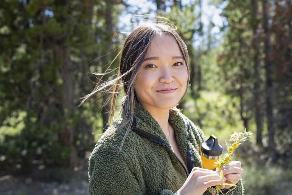 Person in a green jacket holding flowers outdoors, smiling with a forest background.