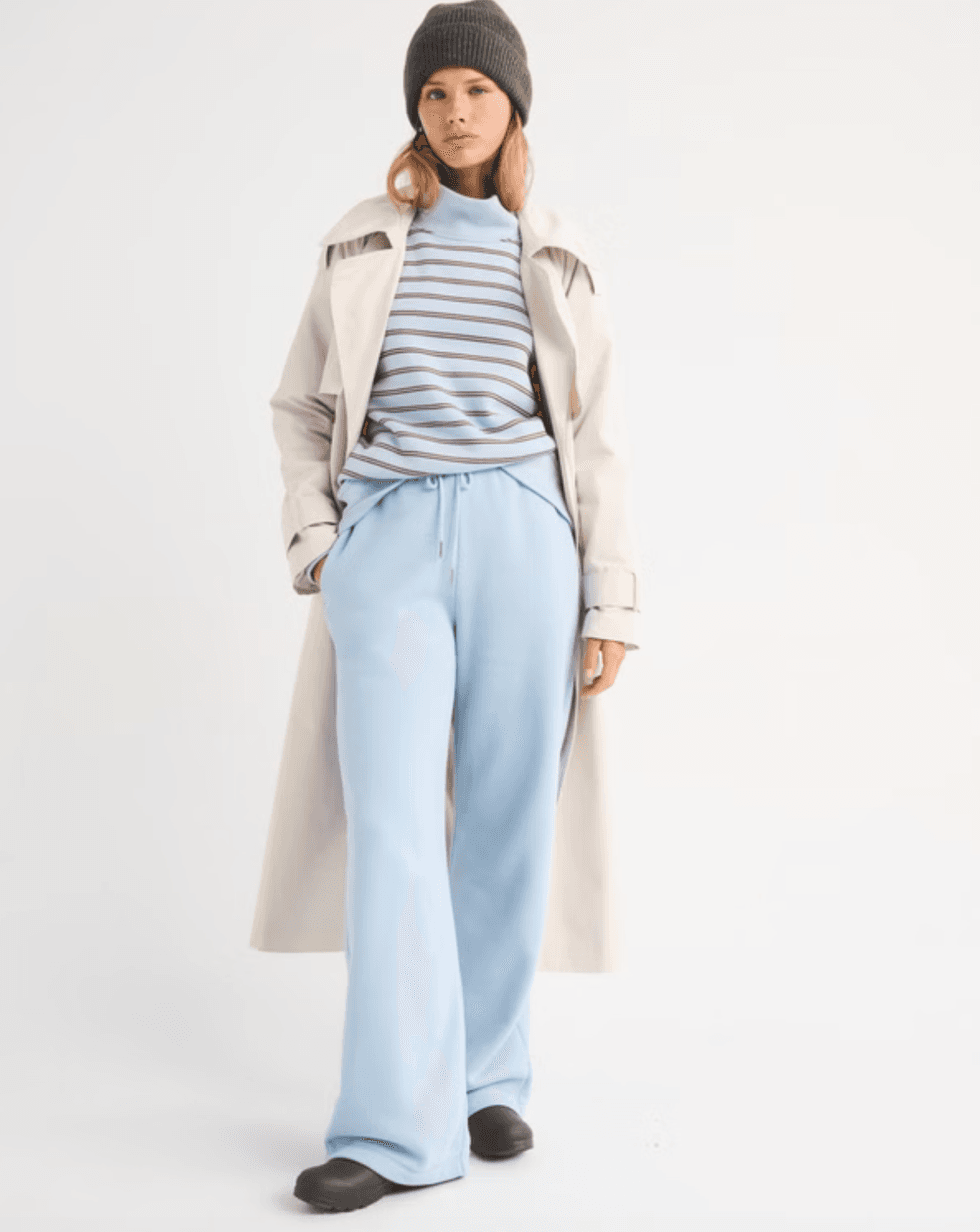 Person in a long coat, striped sweater, light blue pants, and a beanie stands against a white background.