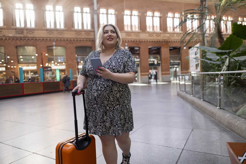 Person in a patterned dress holding phone, with suitcase in a bright, airy hall.