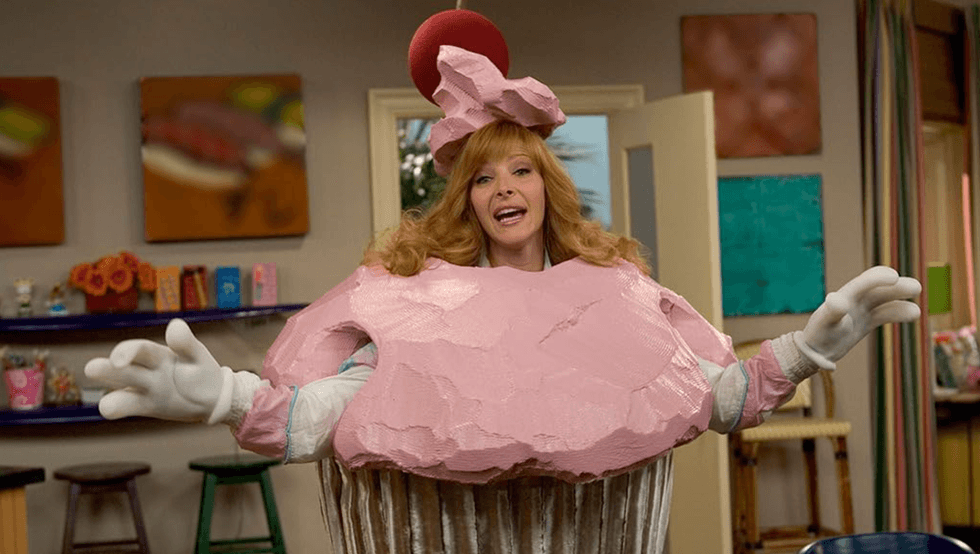 Person in a pink cupcake costume with a cherry hat, in a colorful room.