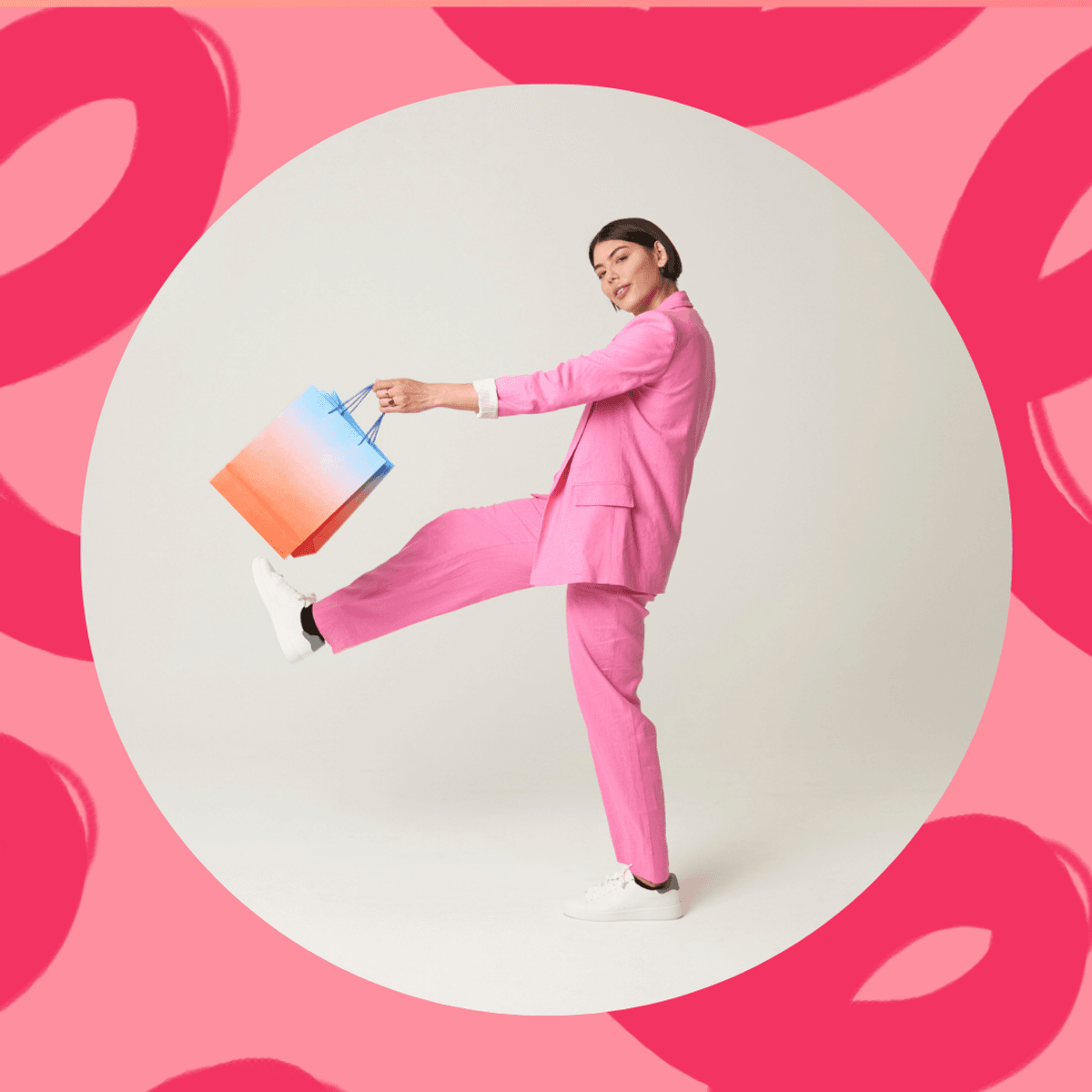 Person in a pink suit holding a colorful shopping bag, posing playfully on a pink background.
