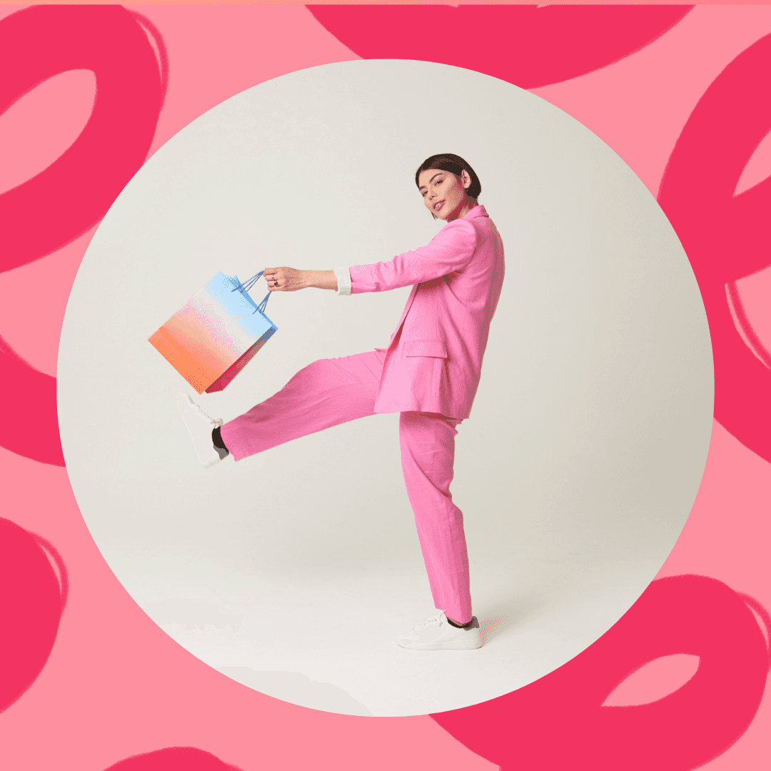 Person in a pink suit holding a colorful shopping bag, posing playfully on a pink background.