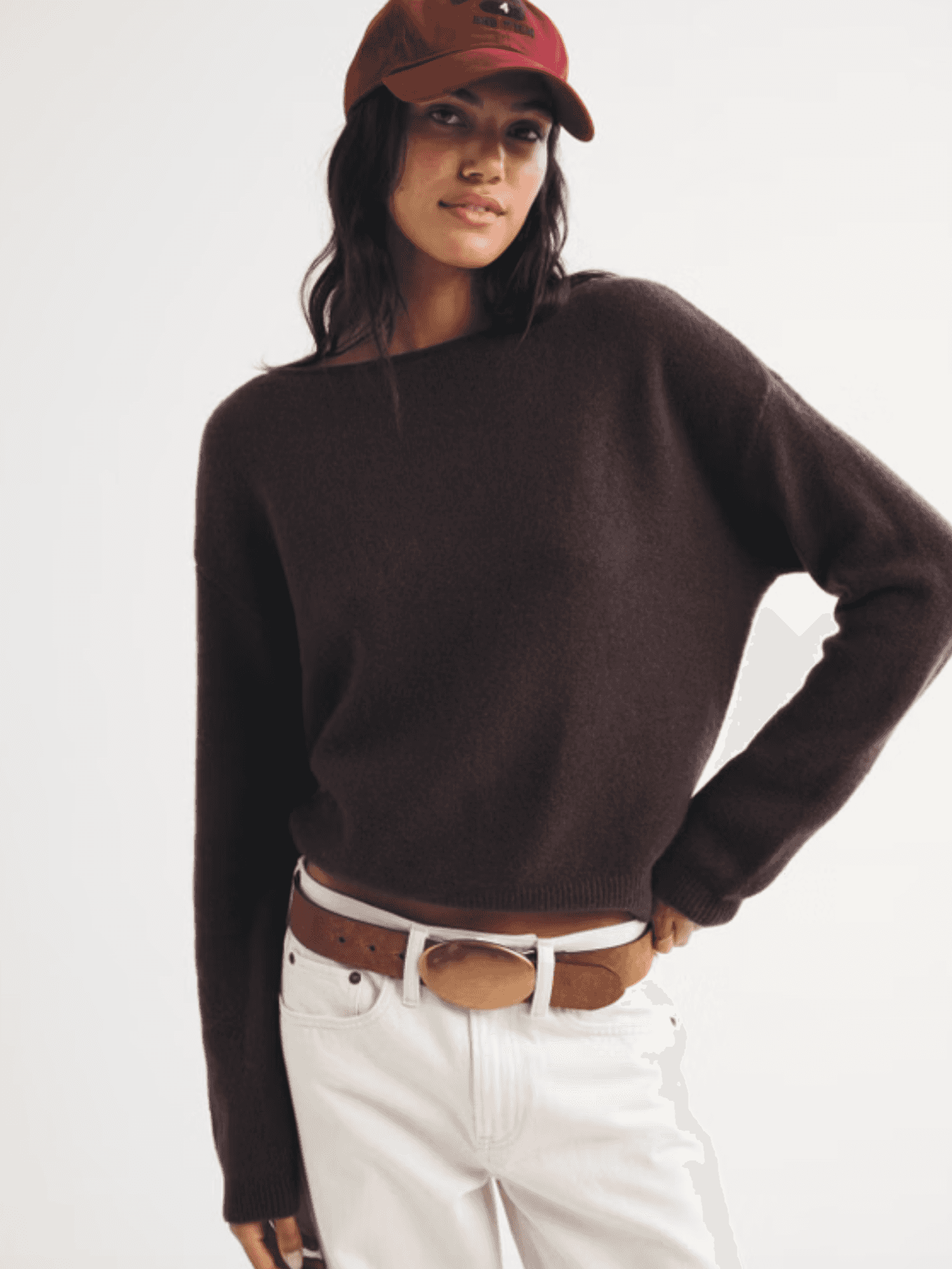 Person in a red cap, brown sweater, and white pants with hands on hips, posing confidently.