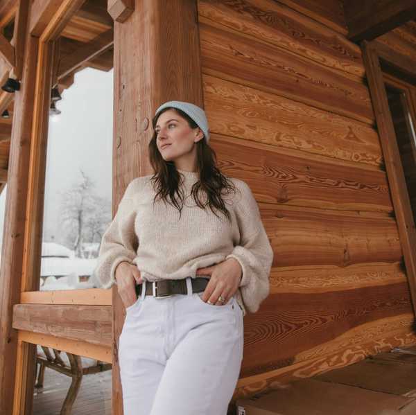 Person in a sweater and beanie, leaning on a wooden cabin railing, with snowy scenery behind.