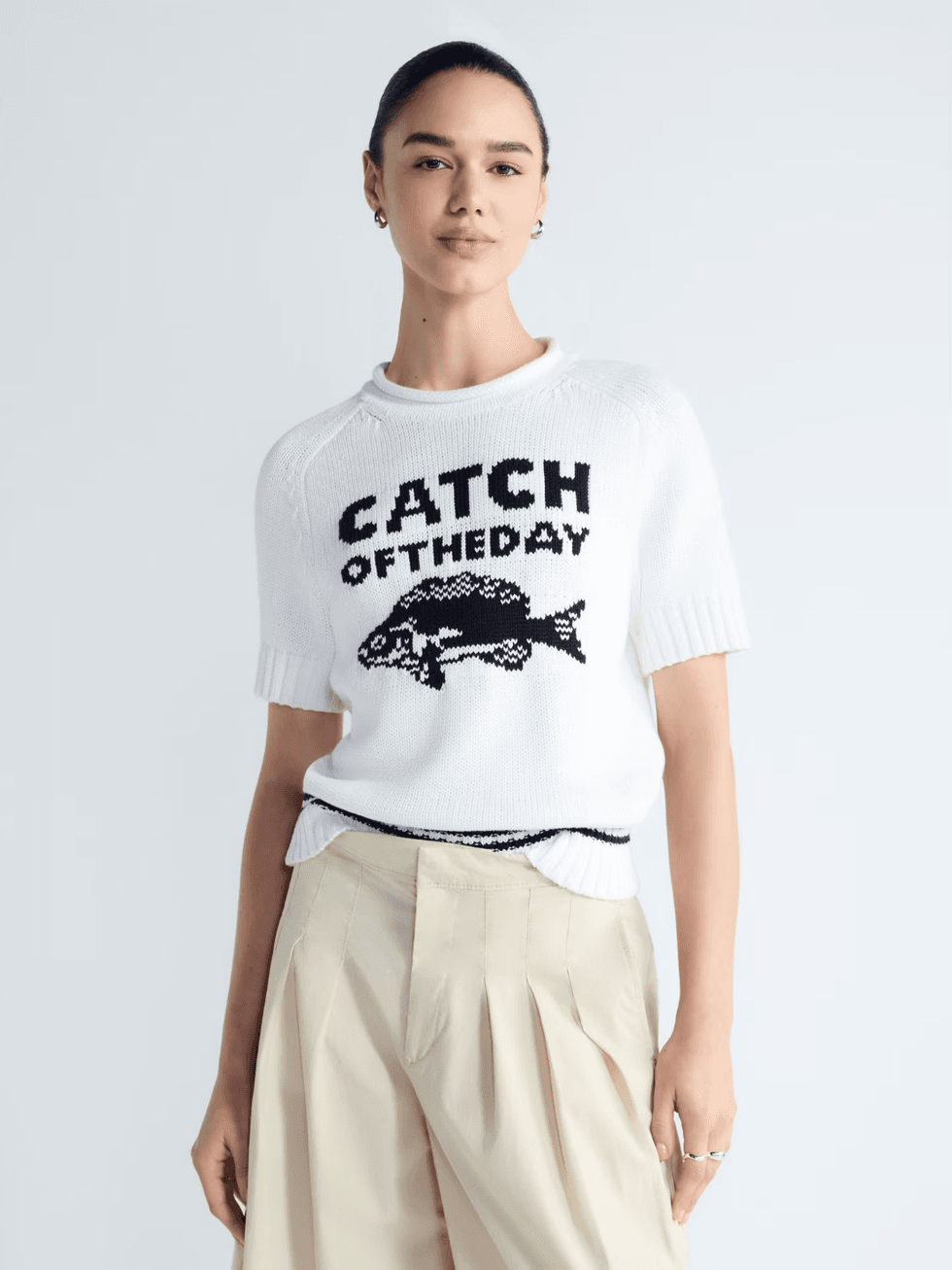 Person in a white "Catch of the Day" sweater with fish design, wearing beige pants.