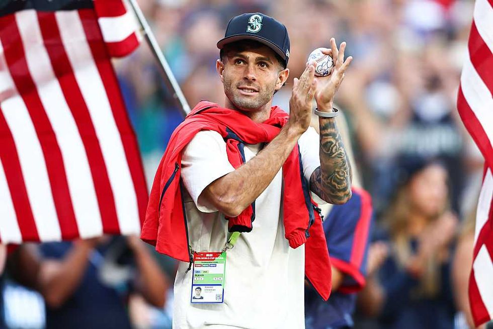 Person in baseball cap holds a ball, American flags in background.