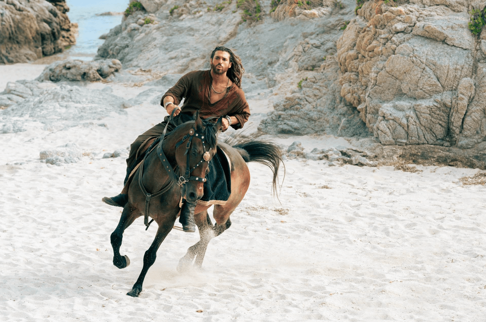 Person in brown clothing riding a horse on a sandy beach with rocky cliffs.