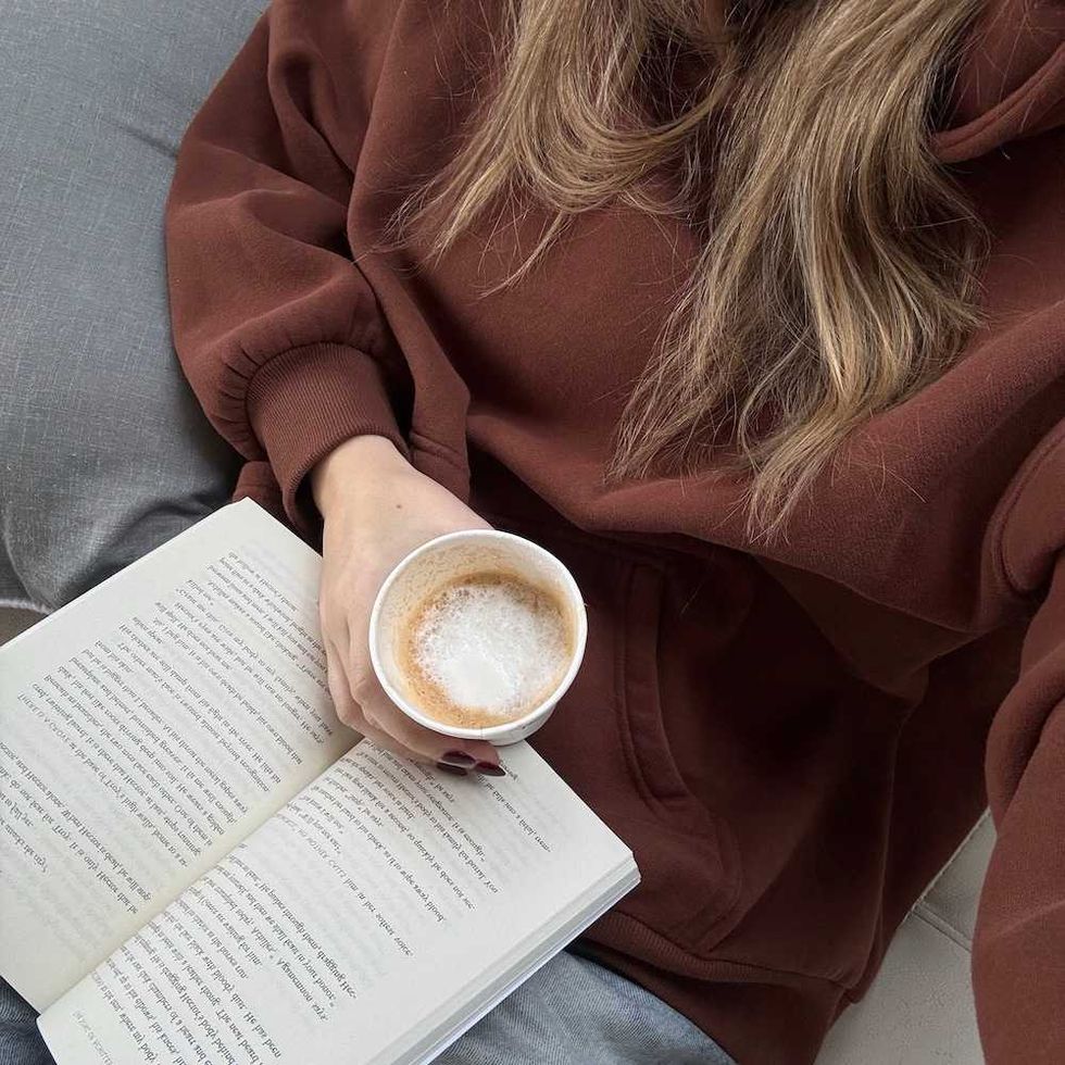 Person in brown sweater reading a book and holding a cup of coffee.