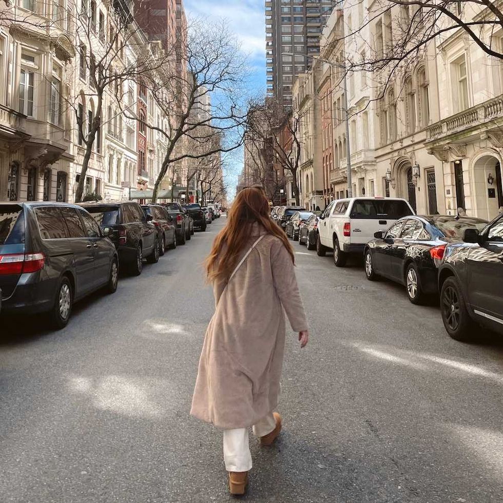 Person in coat walks down a sunny city street lined with cars and tall buildings.