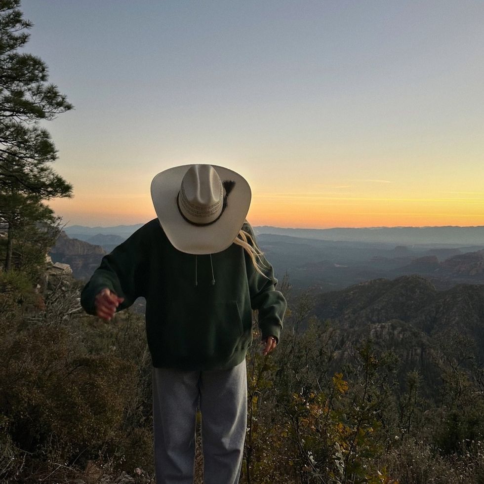 Person in cowboy hat and hoodie in front of scenic mountain sunset.