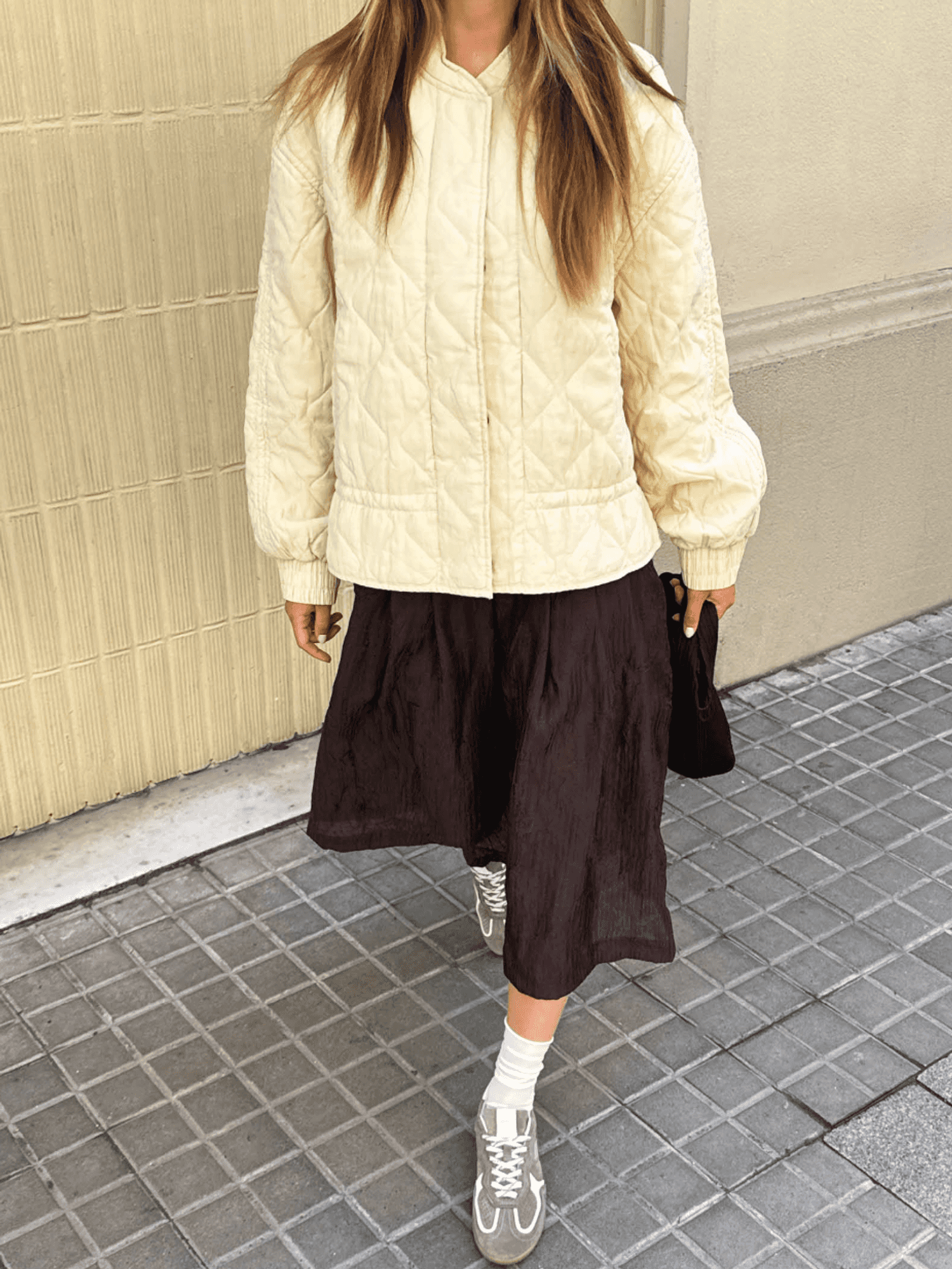 Person in cream quilted jacket, black skirt, gray sneakers on tiled sidewalk.