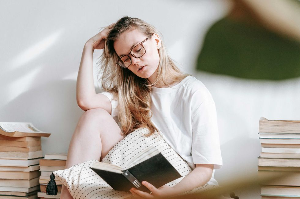 Person in glasses reading a book, surrounded by stacks of books, in a softly lit room.
