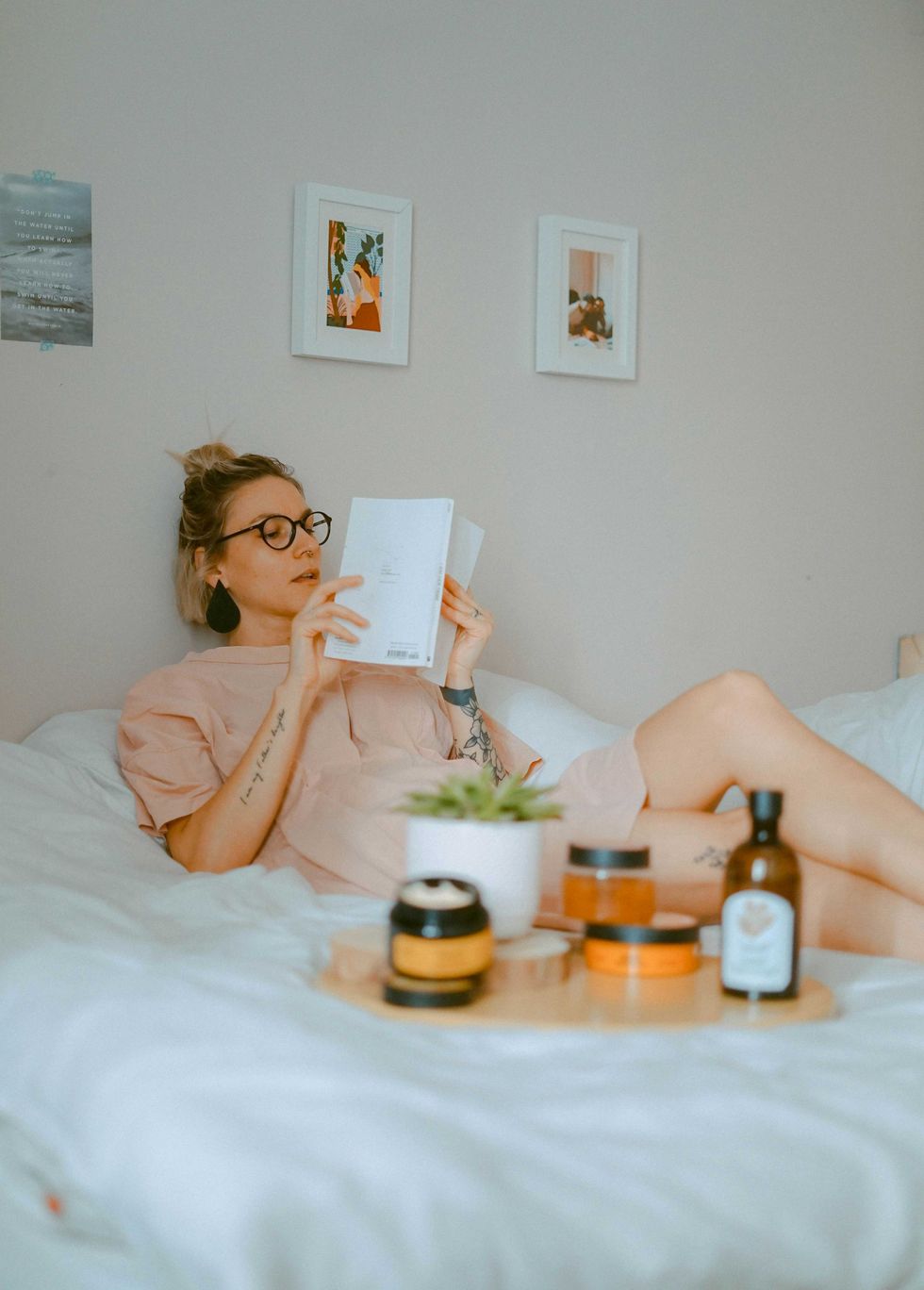 Person in glasses reading on a bed, with skincare items and plant nearby.