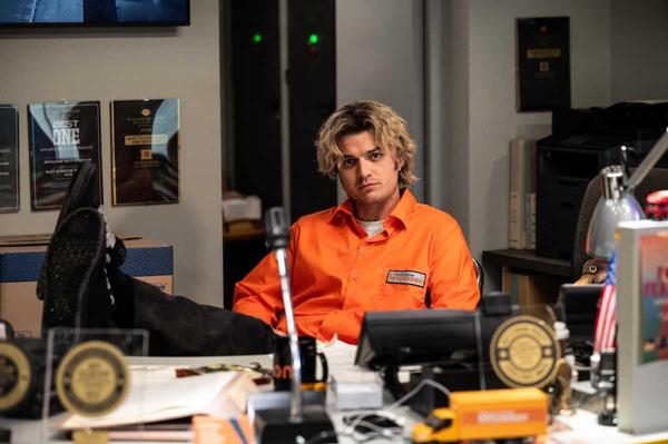 Person in orange uniform sits at cluttered desk with feet up, looking relaxed. joe keery movie