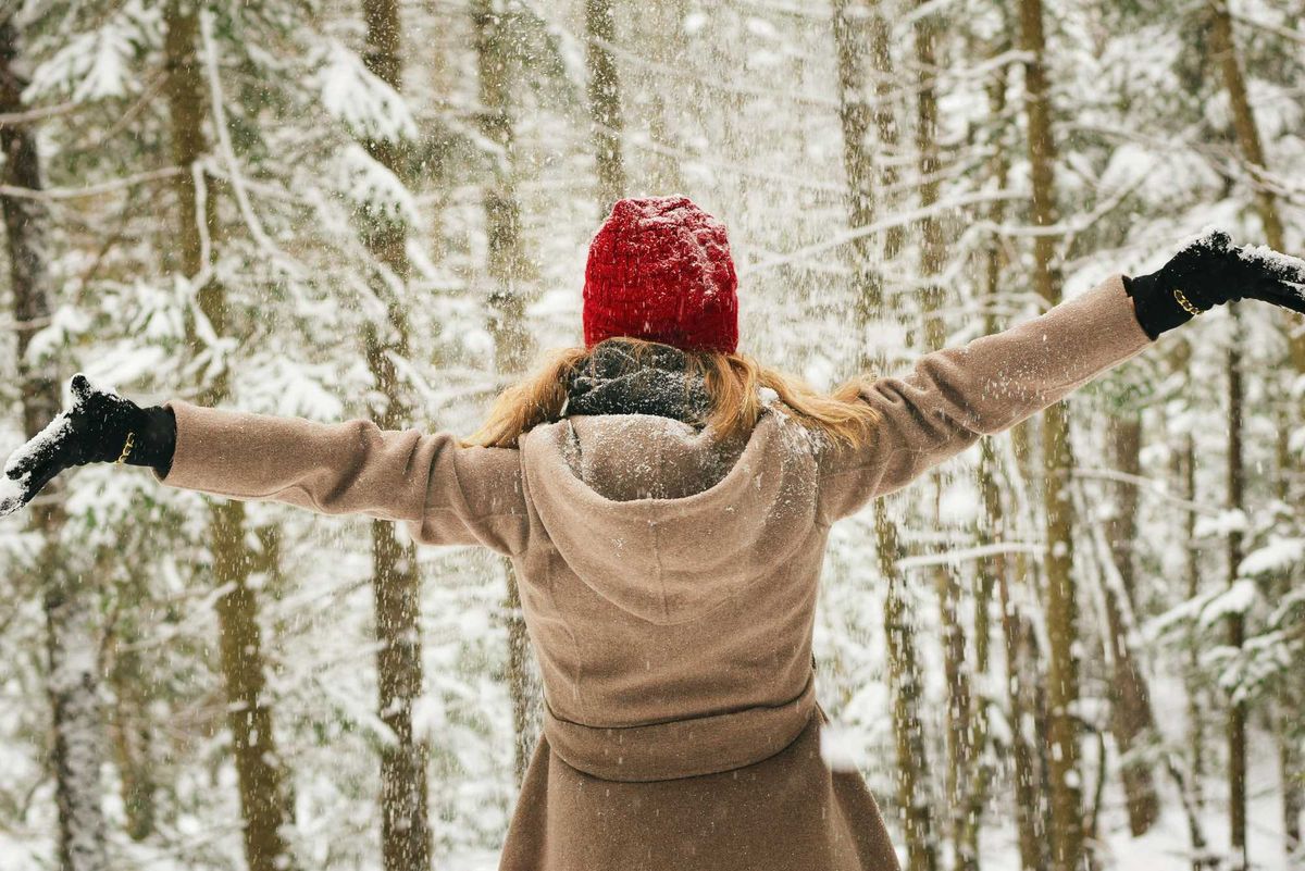 Person in red hat enjoying snowfall in a snowy forest with arms outstretched.