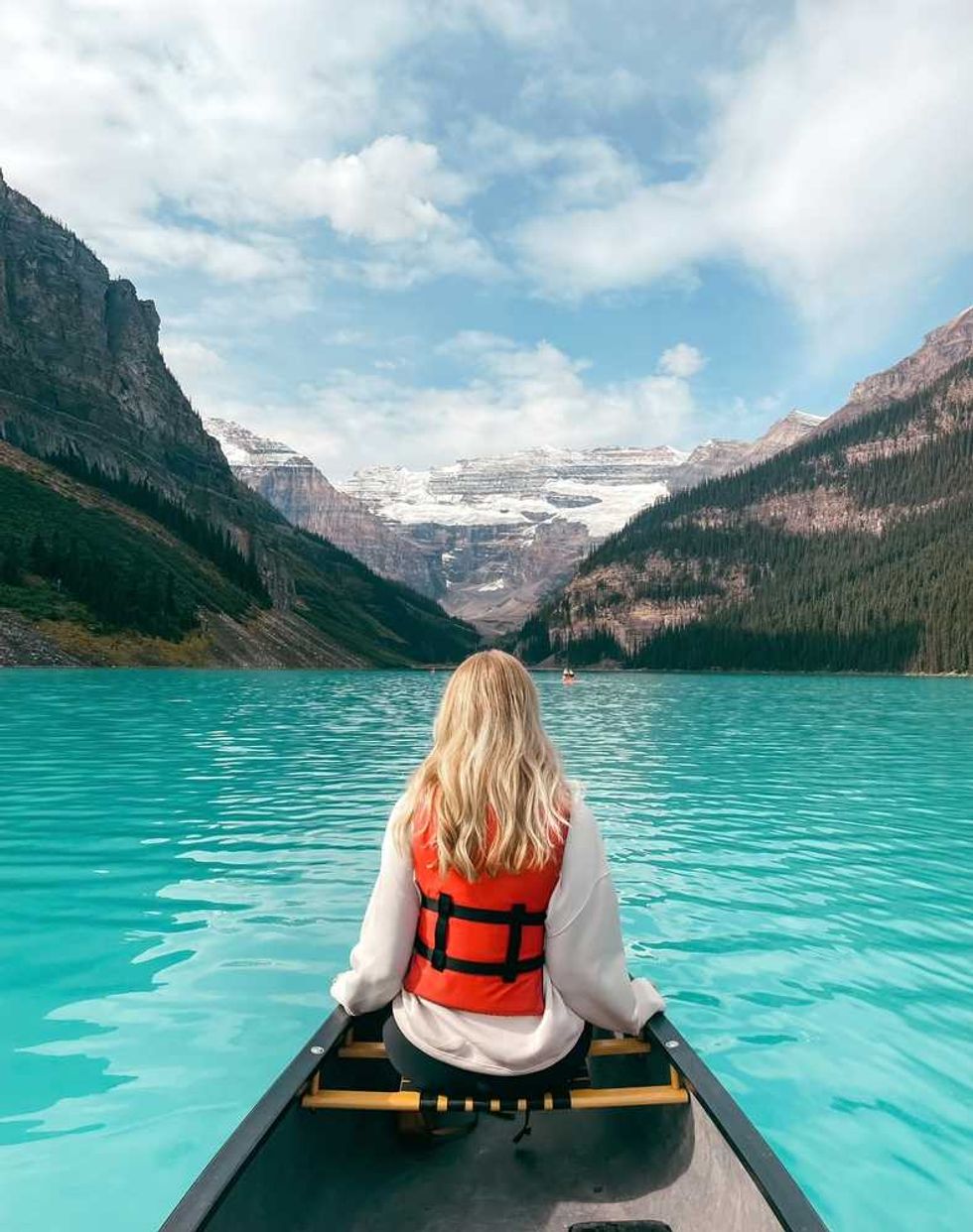 Person in red life jacket canoeing on turquoise lake surrounded by mountains.