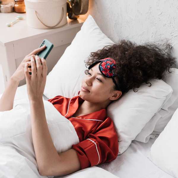 Person in red pajamas on bed, using a phone, wearing a floral sleep mask.