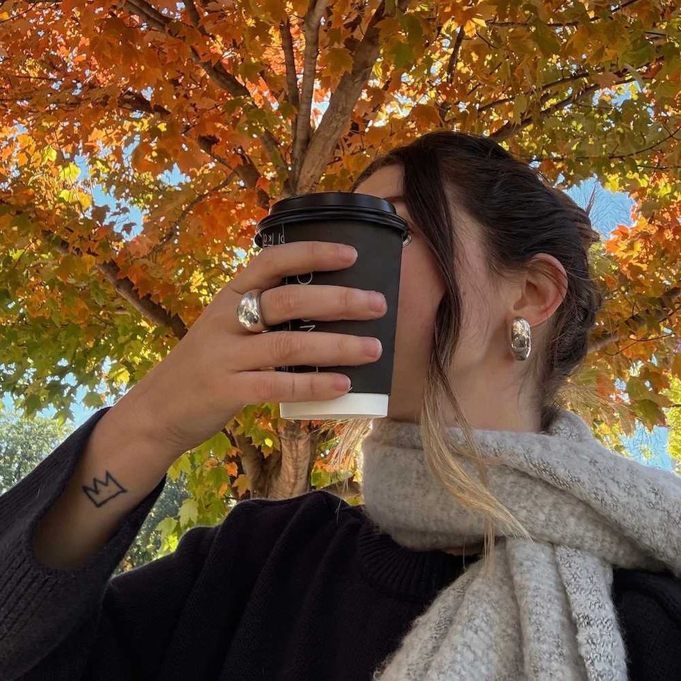 Person in scarf sipping coffee beneath autumn leaves.