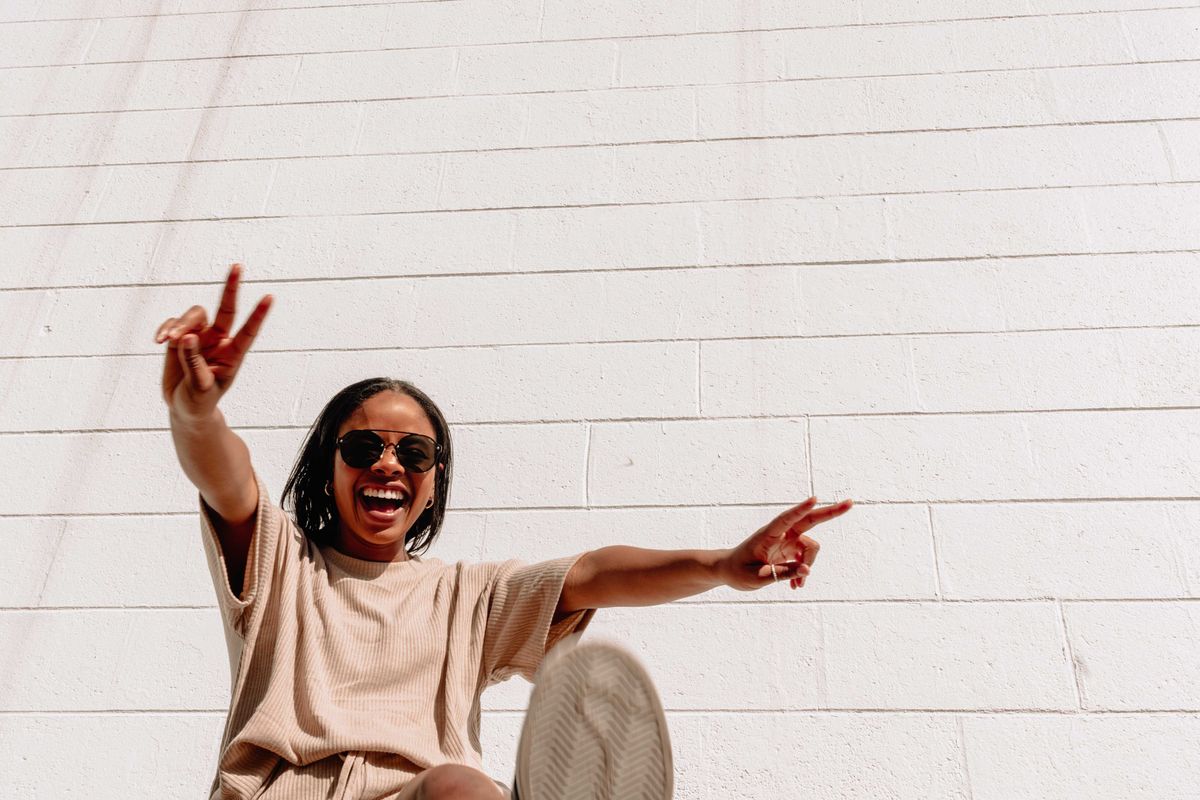 Person in sunglasses smiling, making peace signs against a white brick wall.