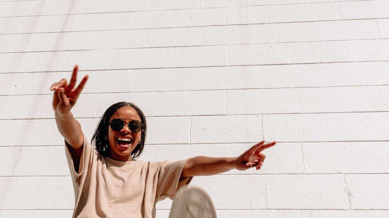 Person in sunglasses smiling, making peace signs against a white brick wall.