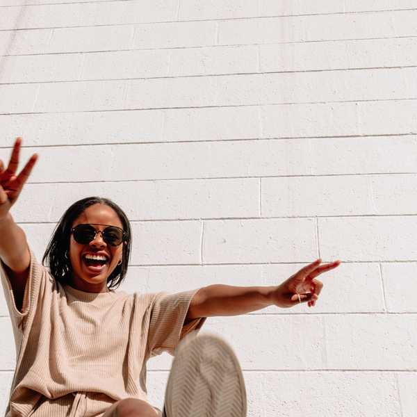 Person in sunglasses smiling, making peace signs against a white brick wall.