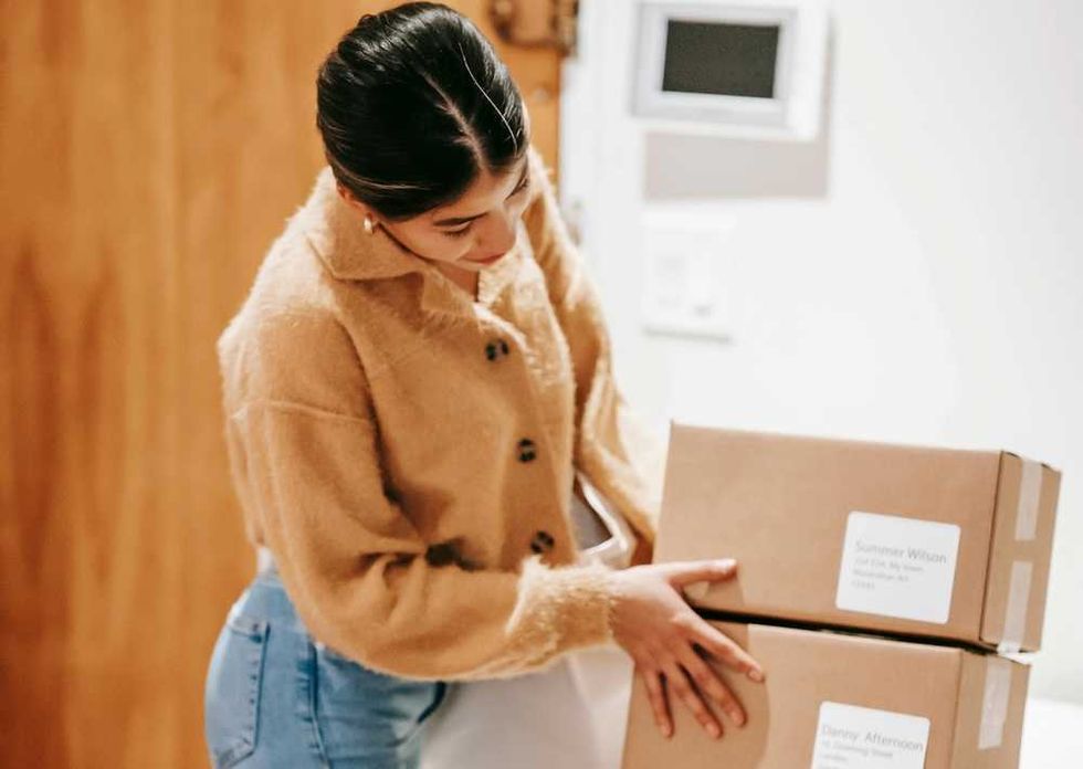 Person in tan sweater holds two cardboard boxes indoors.