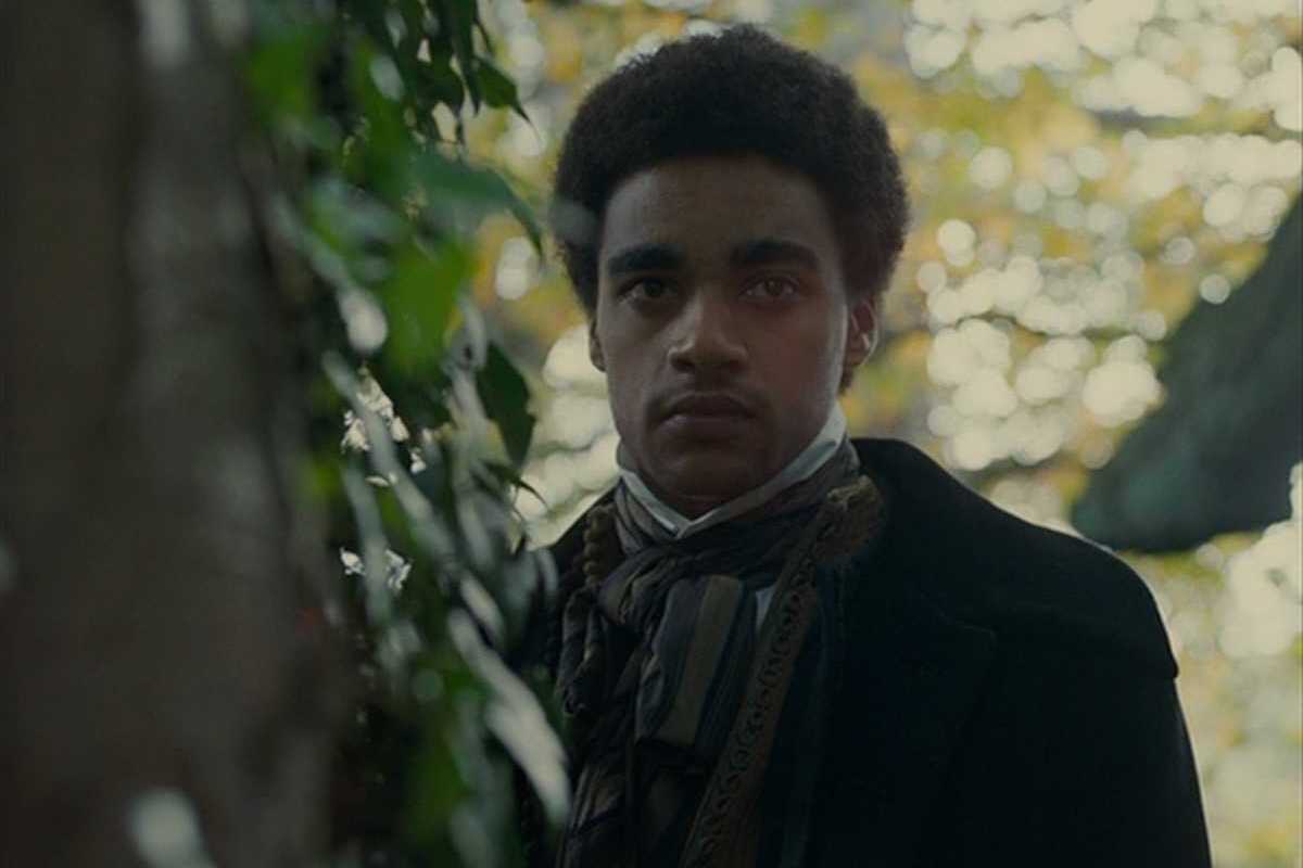 Person in vintage attire stands beside foliage, looking forward.