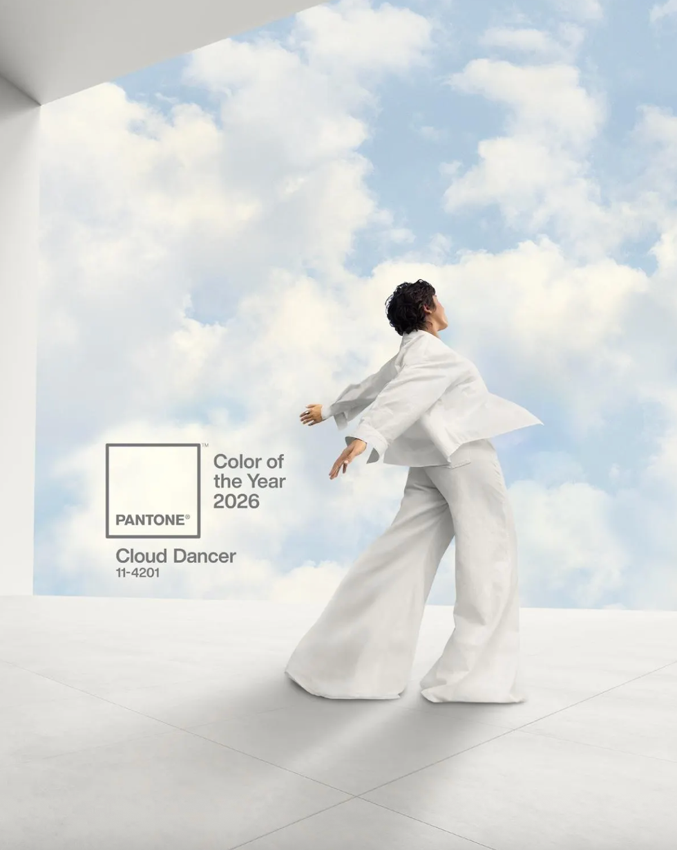 Person in white outfit poses against a cloudy sky, promoting Pantone's 2026 color "Cloud Dancer."