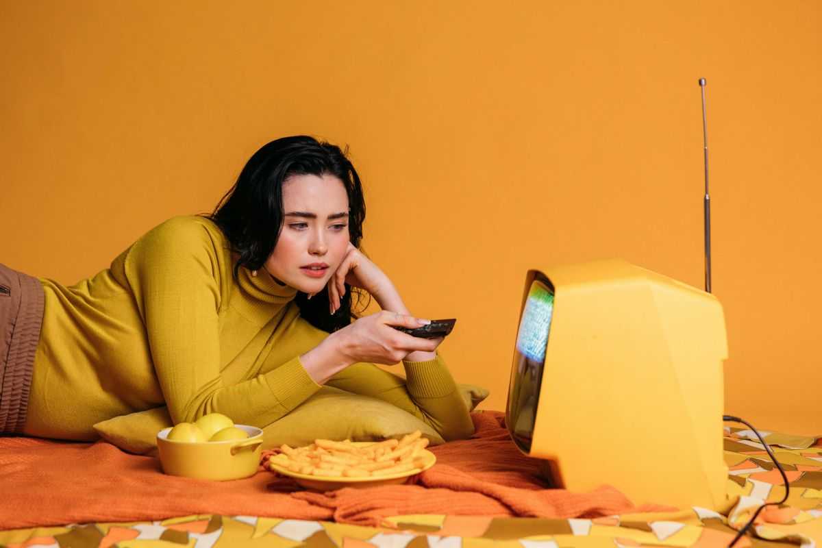 Person in yellow watches vintage TV, holding remote, surrounded by lemons and fries.