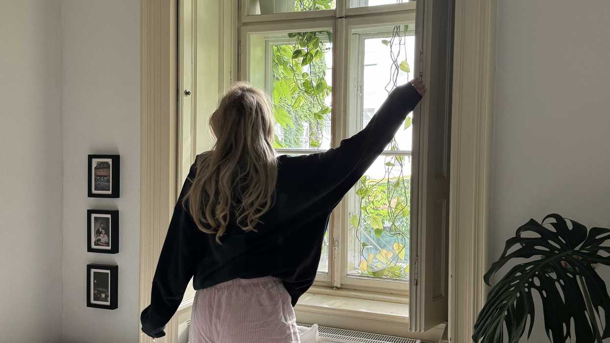 Person kneeling on bed, opening window with greenery outside.