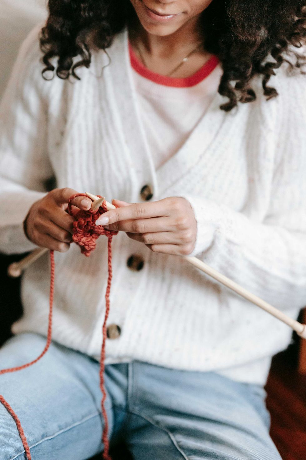 Person knitting with red yarn and wooden needles, wearing a white sweater.