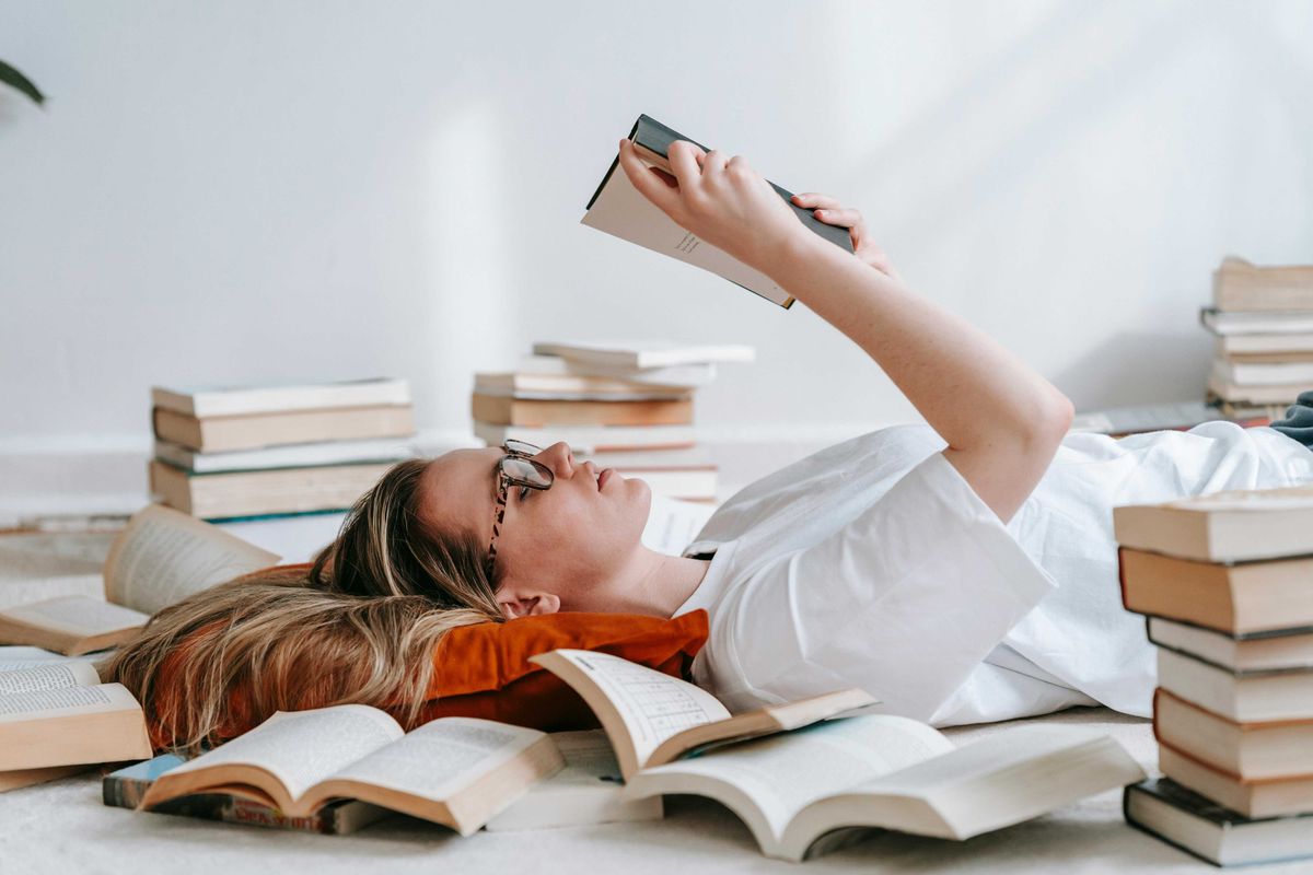 Person lying on the floor, reading a book, surrounded by piles of open and closed books.
