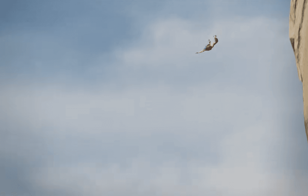 Person mid-air leaping from a rock cliff against a blue sky background.