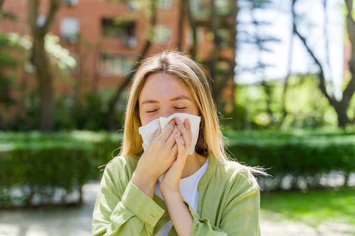 Person outdoors sneezing into a tissue, with green foliage and buildings in the background.