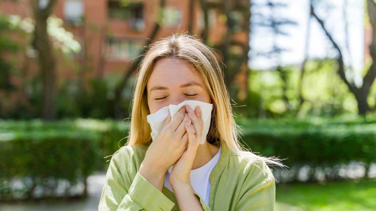 Person outdoors sneezing into a tissue, with green foliage and buildings in the background.