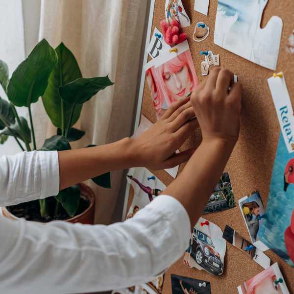 Person pinning images and notes on a corkboard near a plant.