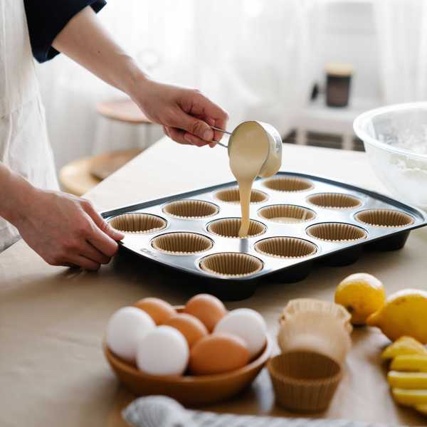 Person pouring batter into cupcake liners on a tray, surrounded by eggs and lemons.