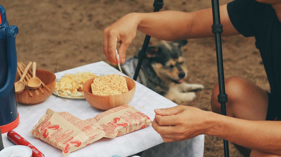 Person preparing instant noodles at an outdoor table with a dog nearby.