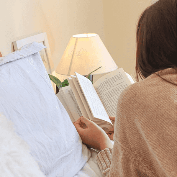 Person reading a book in bed with a lamp nearby, creating a cozy atmosphere.