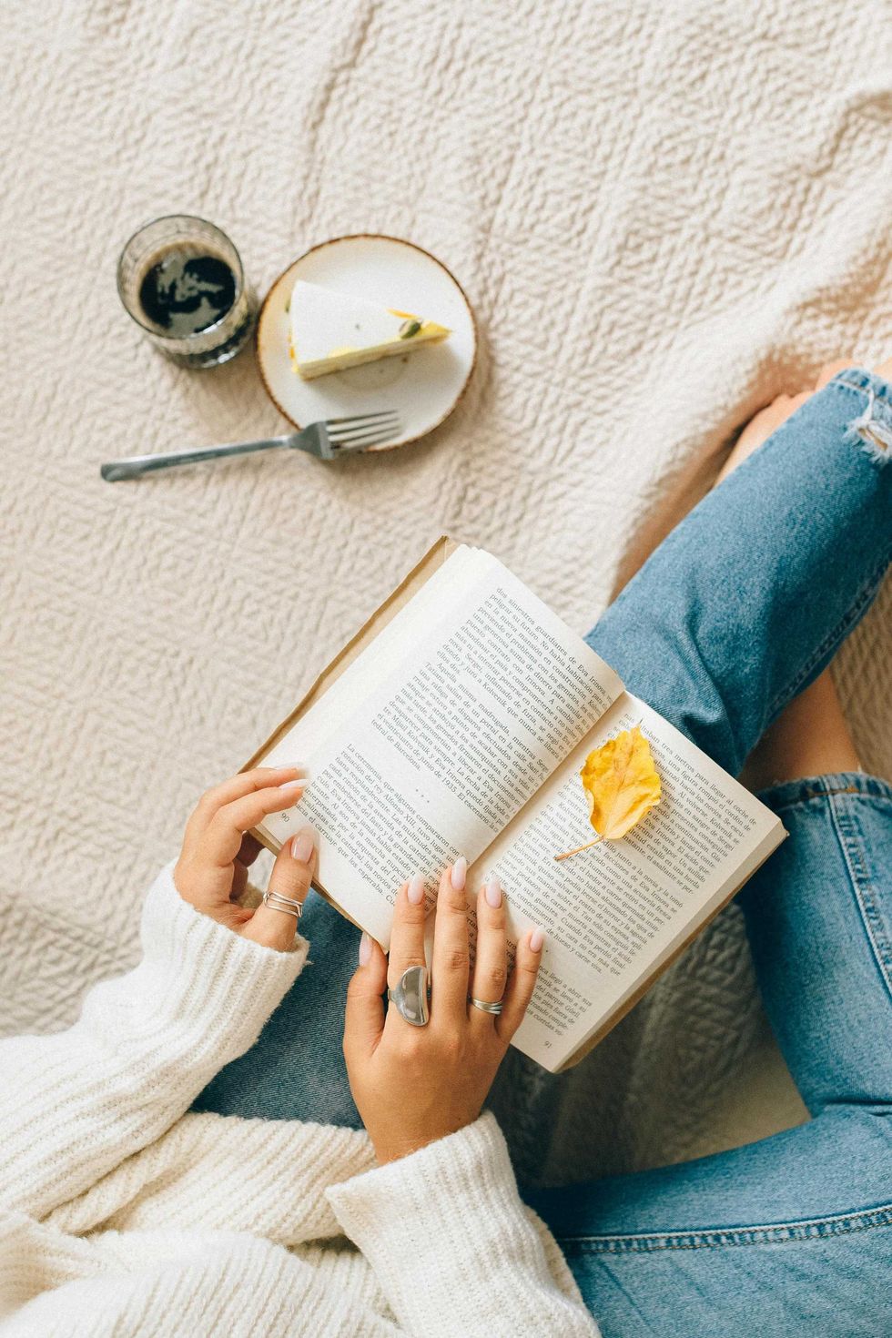 Person reading, holding a book with a yellow leaf; coffee and cake nearby on a knitted blanket.