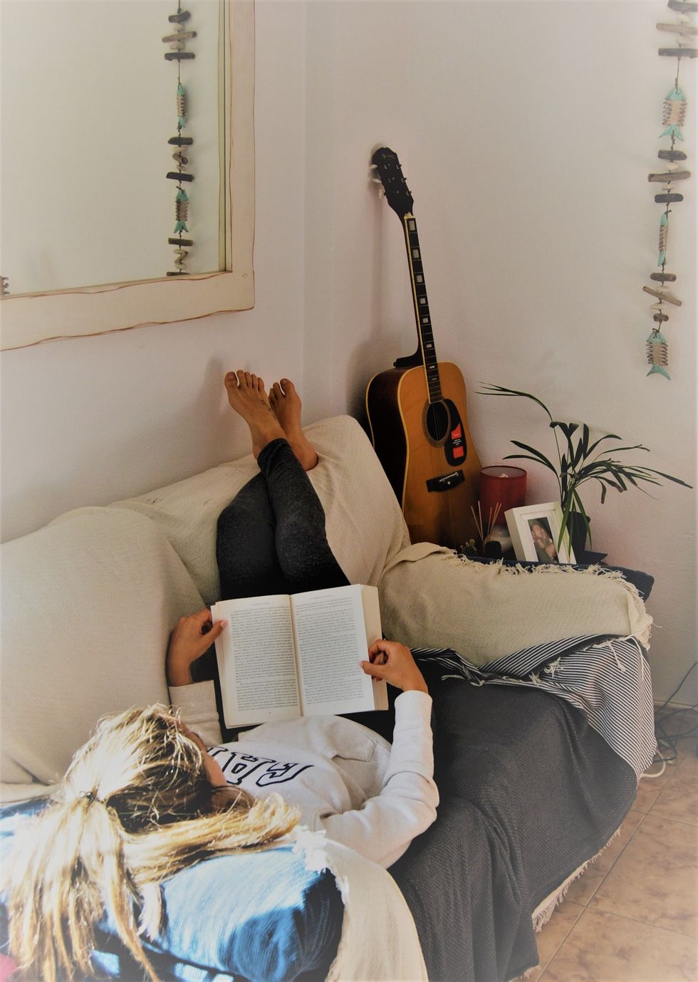 Person reading on a couch with feet up, next to a guitar and indoor plant.